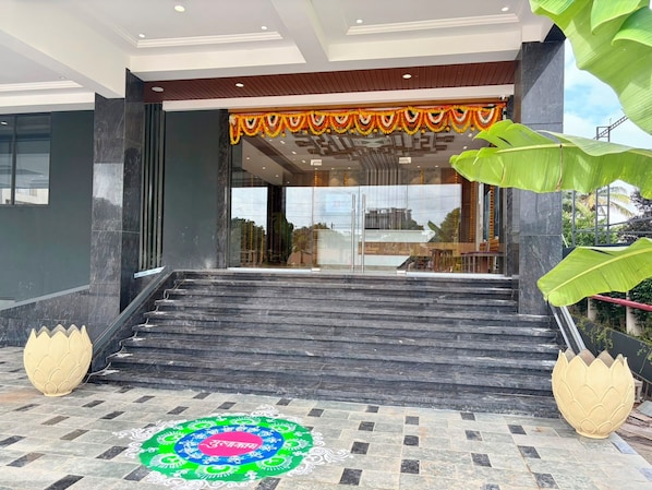 A modern building entrance with a staircase leading to a glass door, a decorative garland above the door, and a traditional rangoli design on the ground.