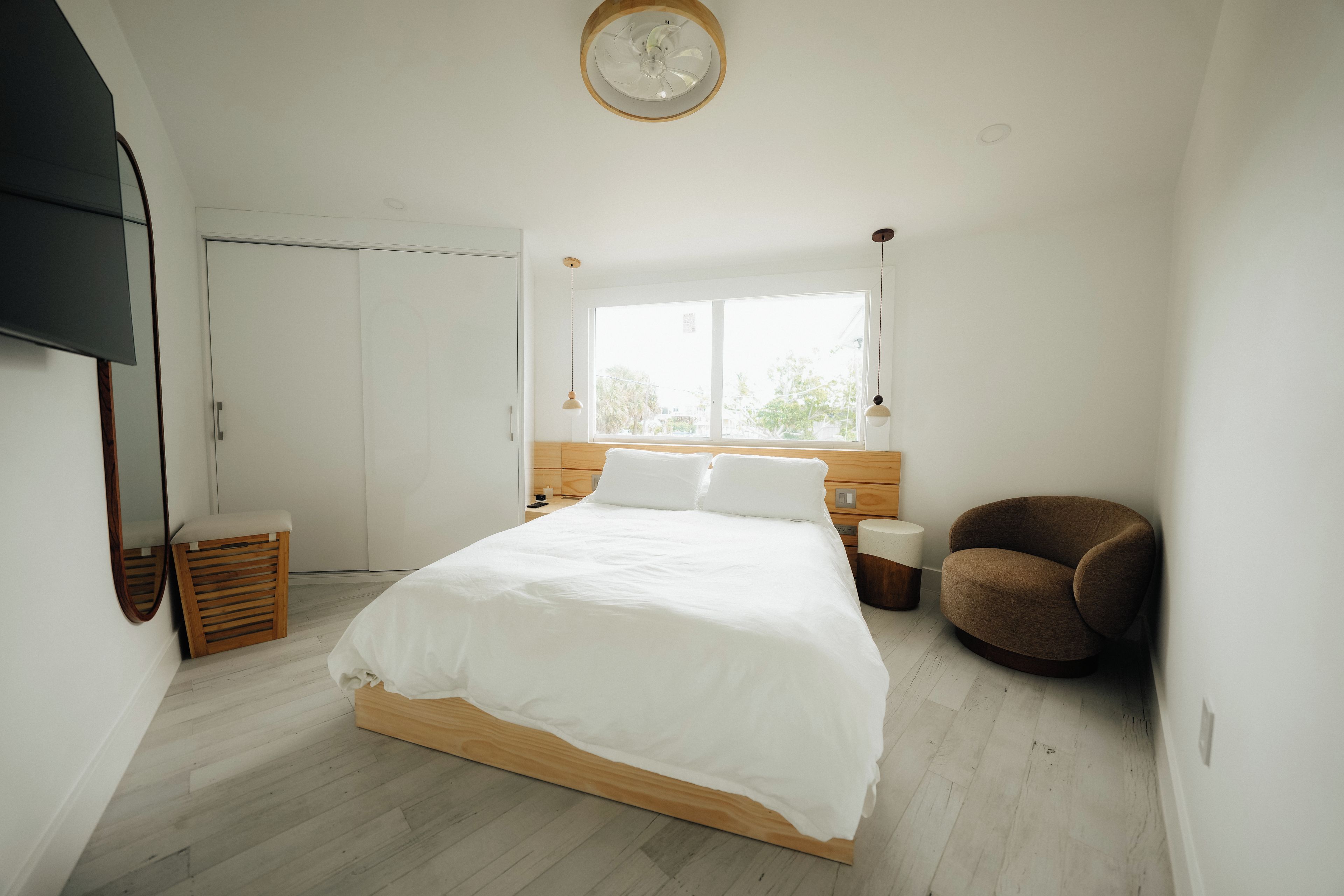 A bedroom with a large bed, a wooden headboard, a bedside table, a chair, a wardrobe, and a window.