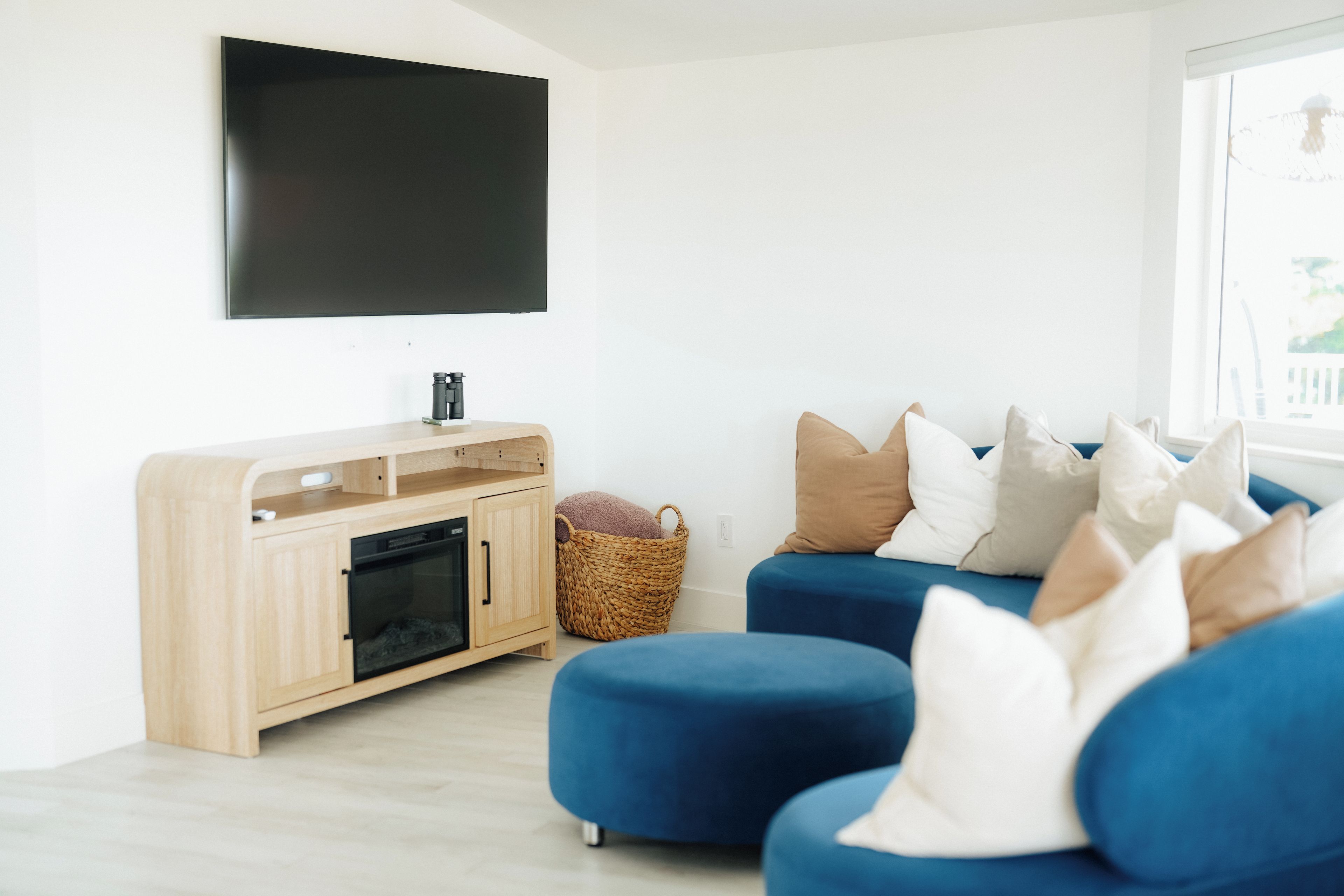 A modern living room with a flat-screen TV mounted on the wall, a wooden entertainment unit with a TV stand, and a blue sofa with multiple cushions.
