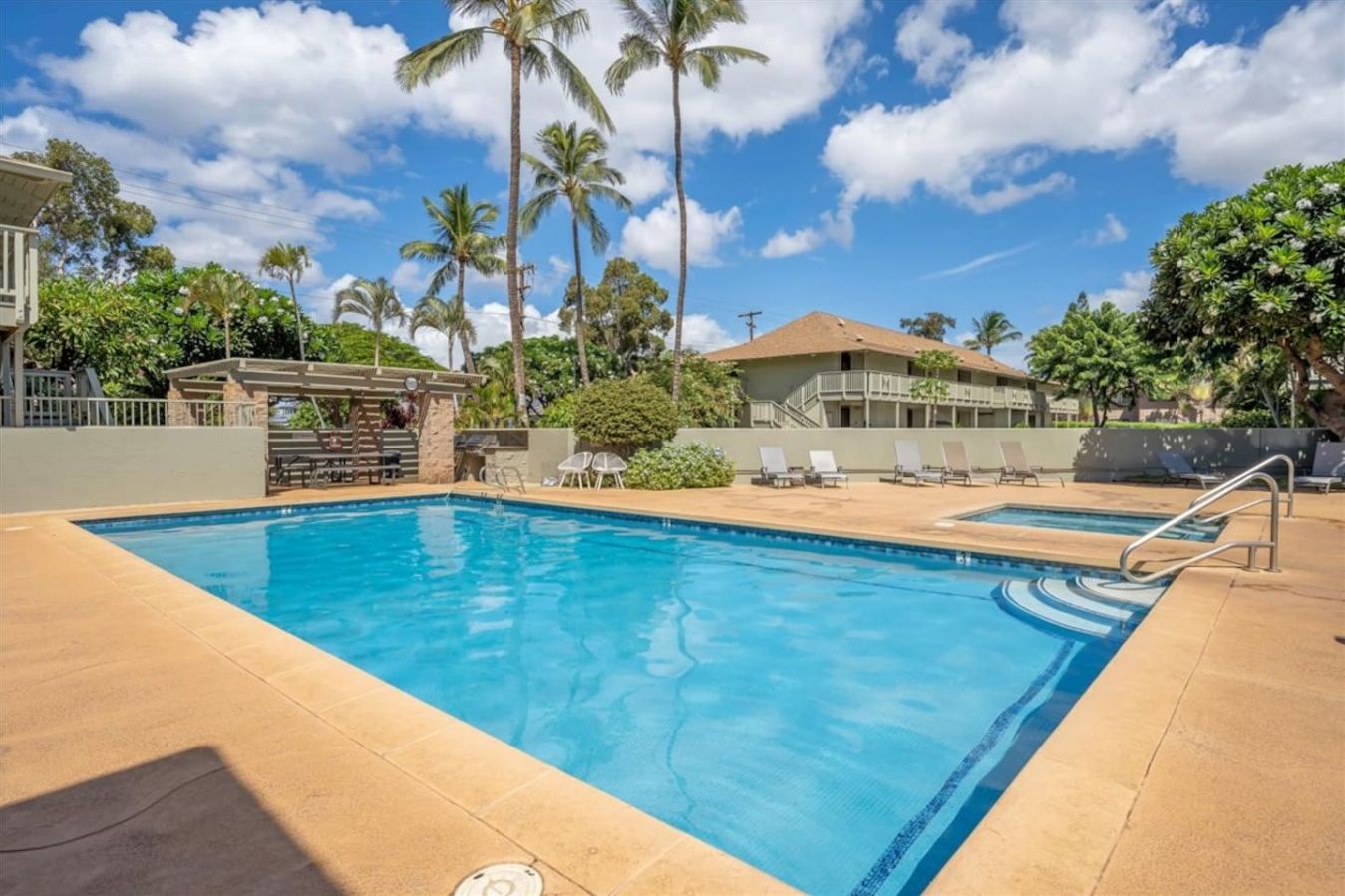 A swimming pool with lounge chairs and a covered seating area, surrounded by palm trees and a building with a balcony.