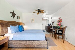 Ein Schlafzimmer mit einem Bett, einem Schreibtisch und einem Stuhl. Es gibt einen Deckenventilator, ein Wandbild und eine kleine Küchenzeile.