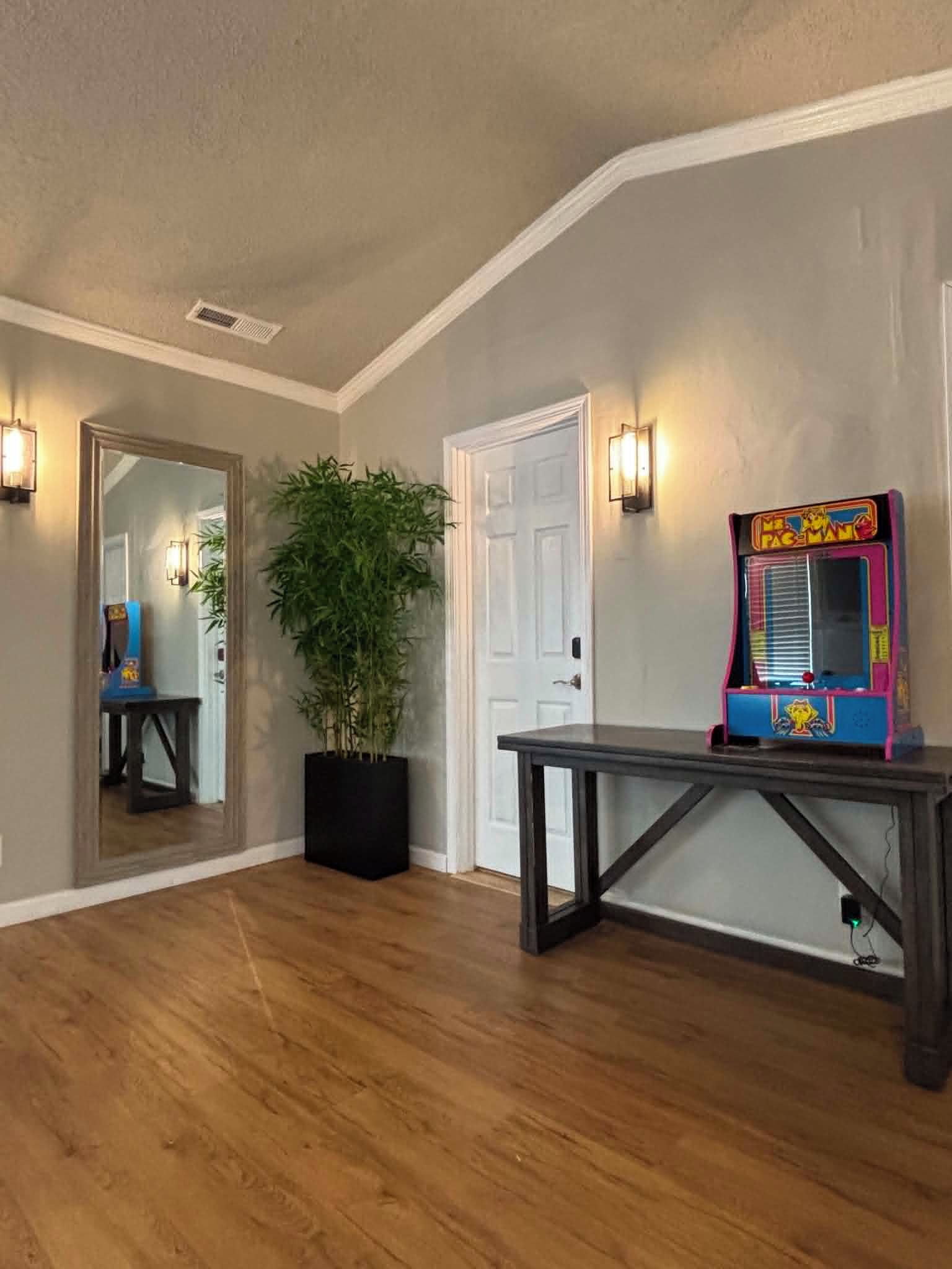 A room with a wooden floor, a potted plant, a table, and a colorful arcade machine.