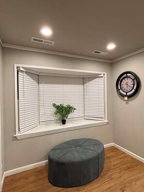 A room with a bay window, a clock, a potted plant, and a grey ottoman.