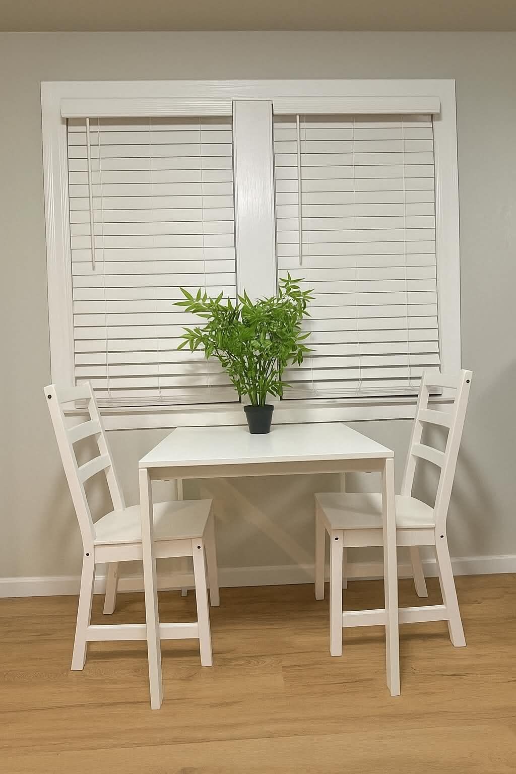 A white table with two chairs and a potted plant in front of a window with blinds.