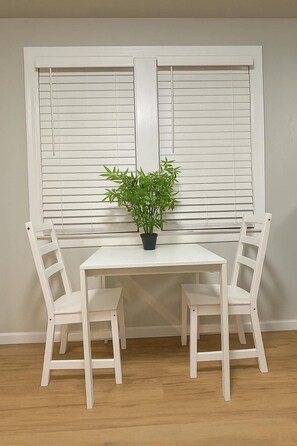 A white table with two chairs and a potted plant in front of a window with blinds.