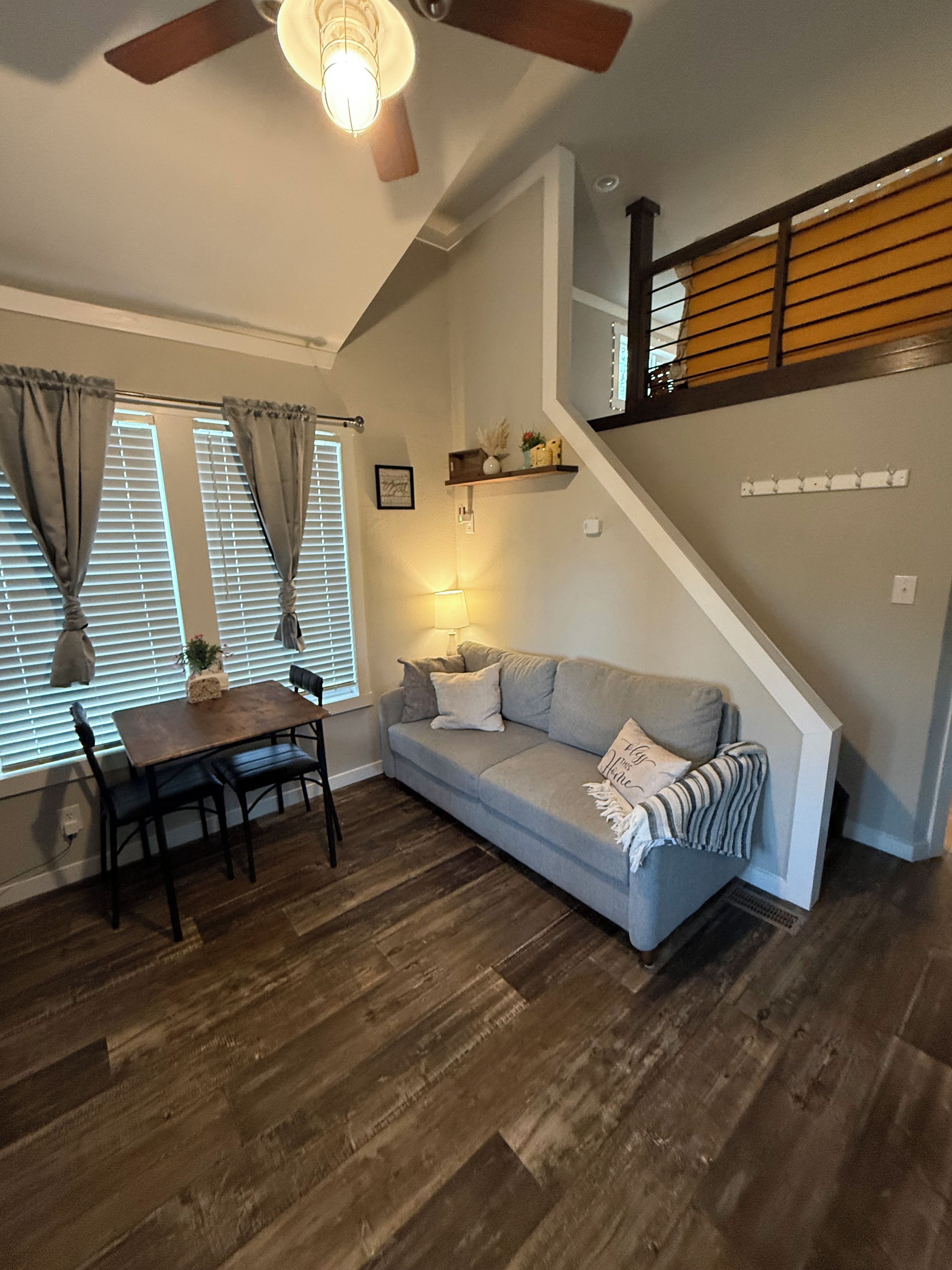 A living room with a sofa, a dining table with chairs, a ceiling fan, and a staircase.