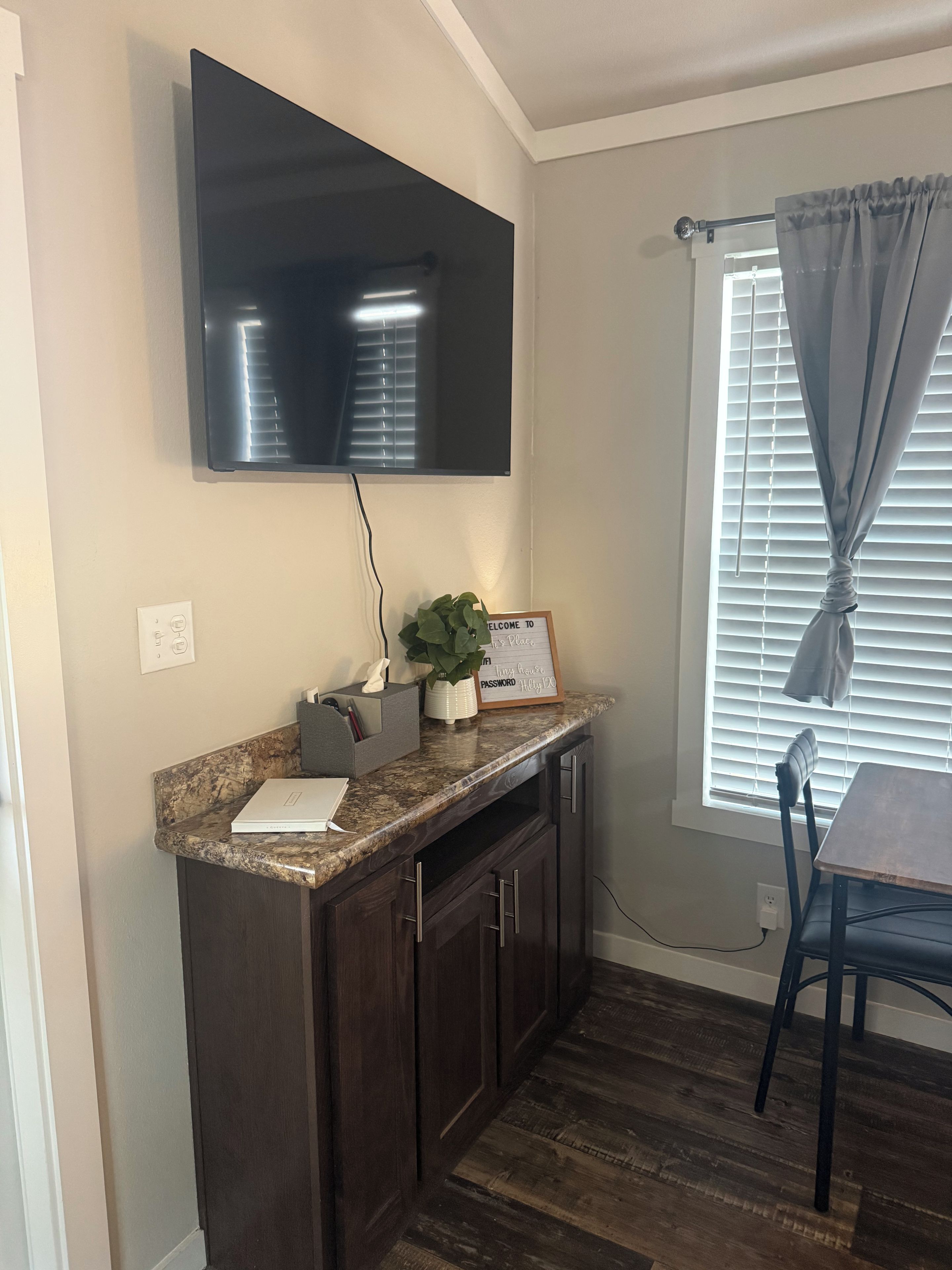 A kitchen with dark wood cabinets, a granite countertop, a flat-screen TV mounted above the counter, a potted plant, and a sign with a message.