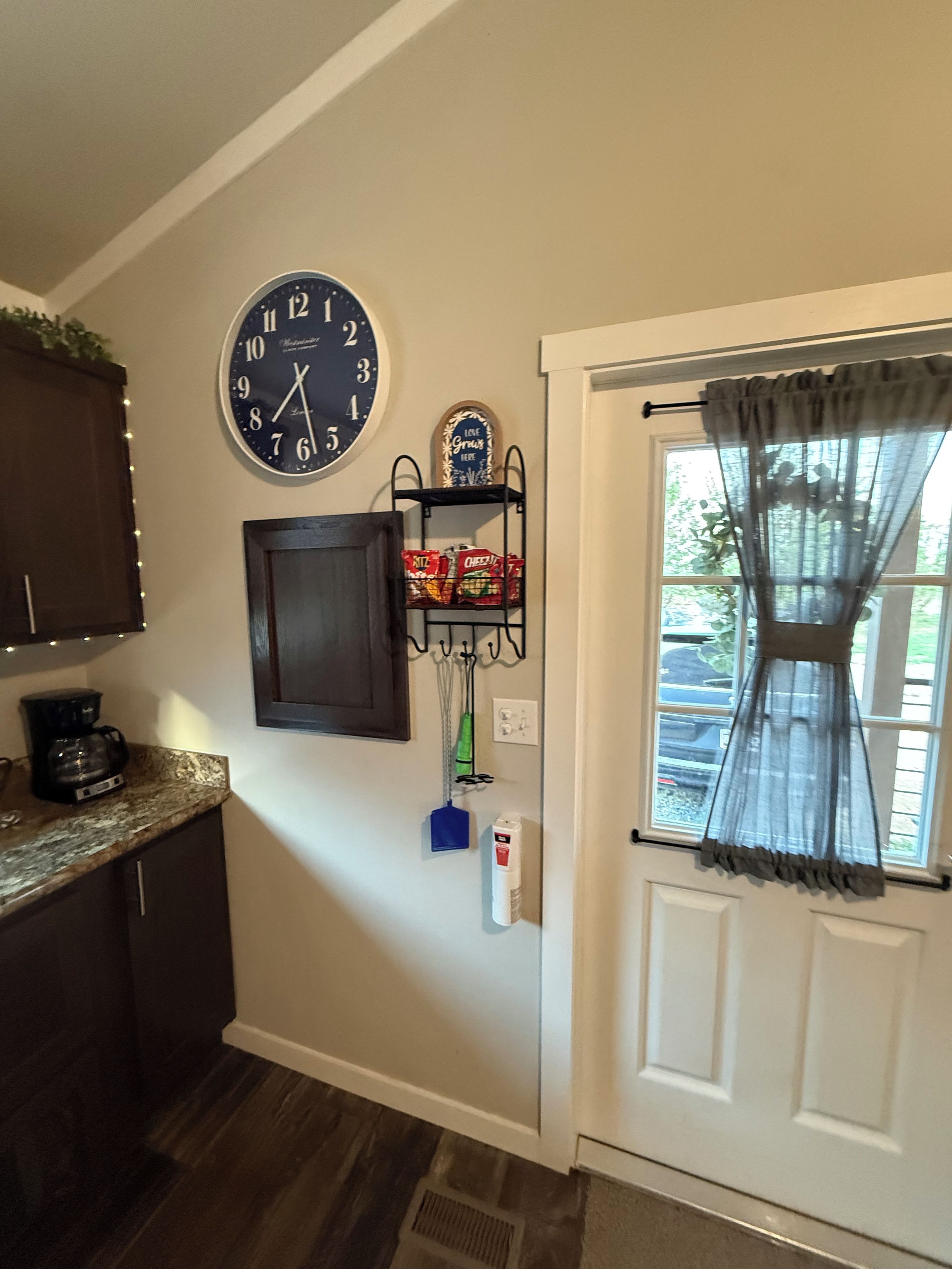 A kitchen with a clock, a coffee maker, a shelf with snacks, and a door with a curtain.
