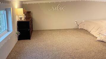 A bedroom with a carpeted floor, a bed, a dresser, a lamp, and a wall sign that says 'relax'.