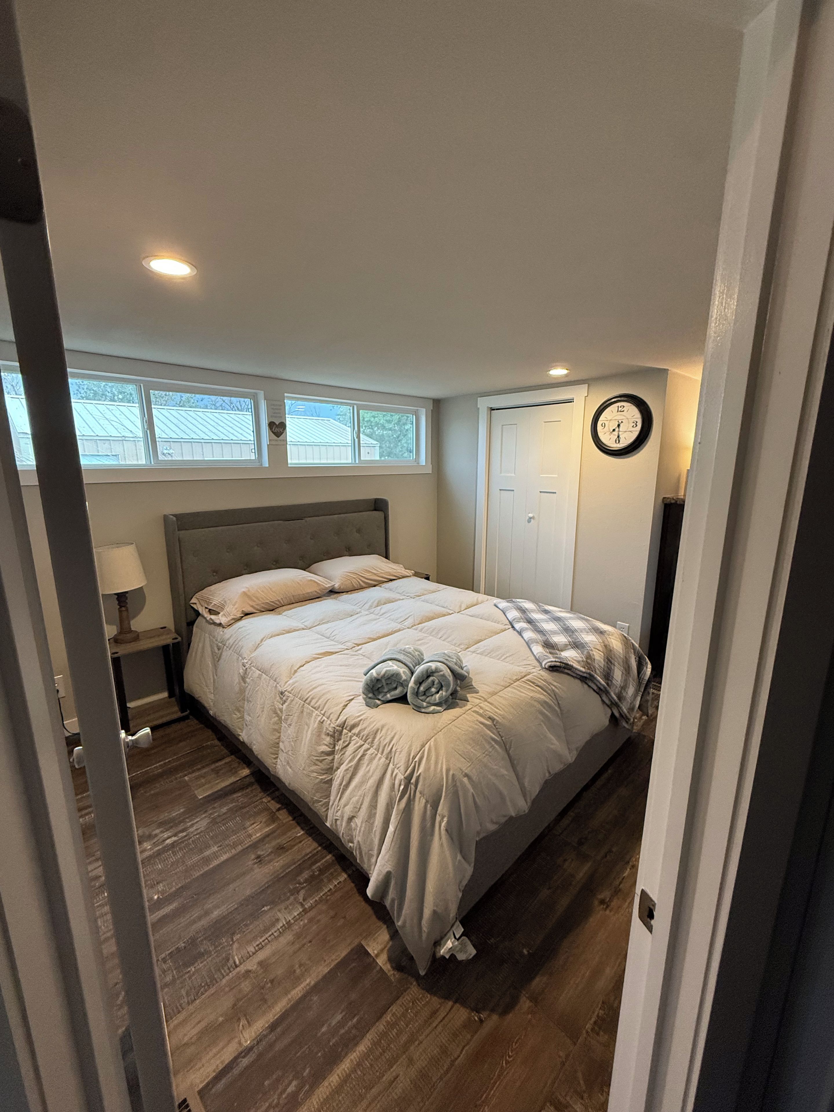 A bedroom with a bed, a nightstand, a lamp, a window, a door, and a wall clock.