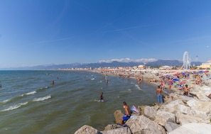 Ein Strand mit badenden und sonnenden Personen, einem Riesenrad und Bergen im Hintergrund.