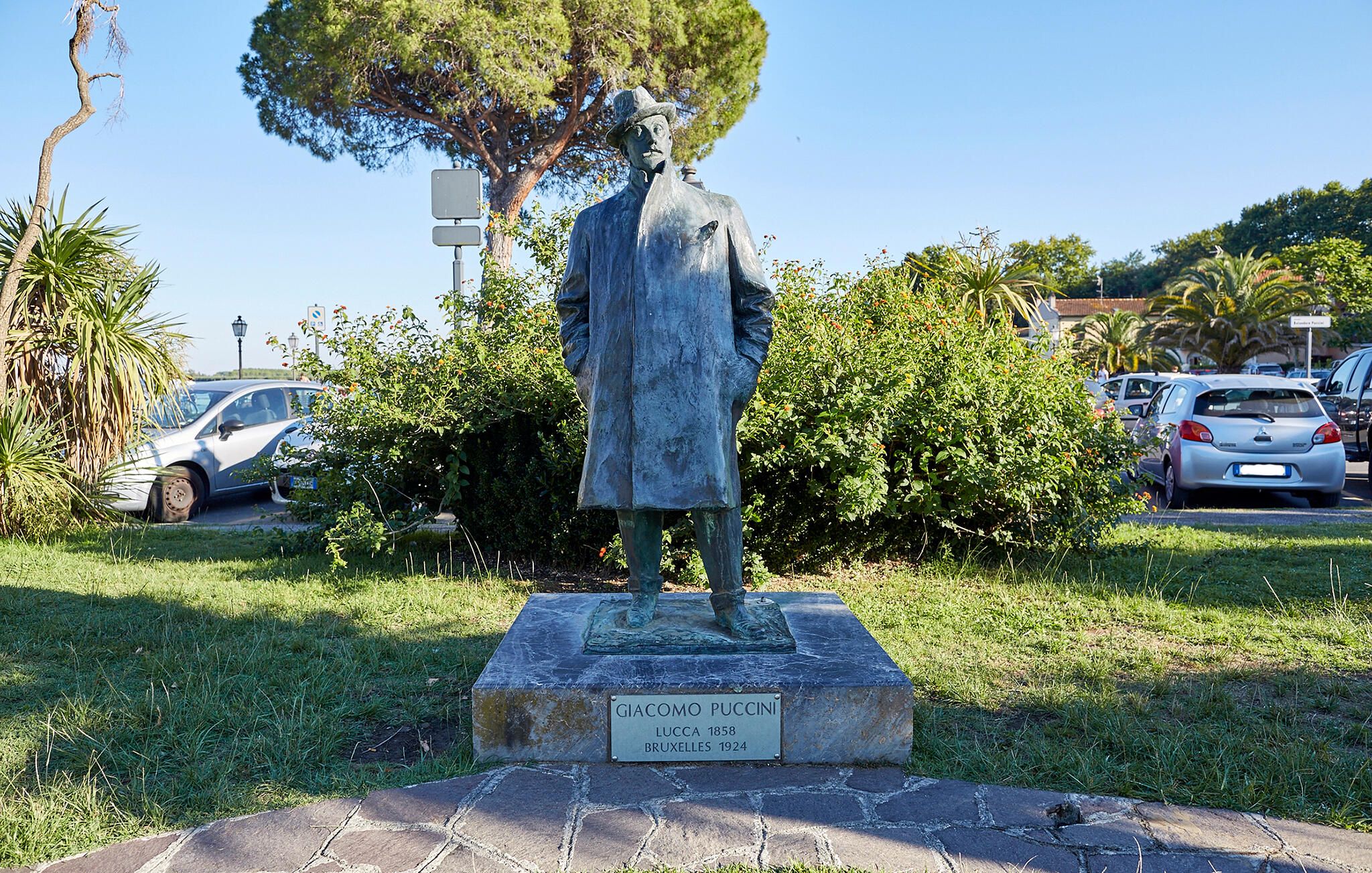 Een standbeeld van Giacomo Puccini in een park, met auto's en bomen op de achtergrond.