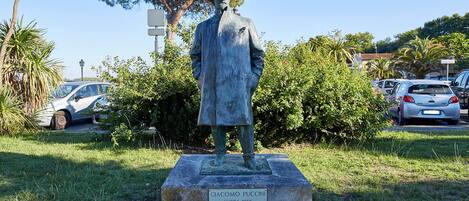 A statue of Giacomo Puccini in a park with cars and trees in the background.