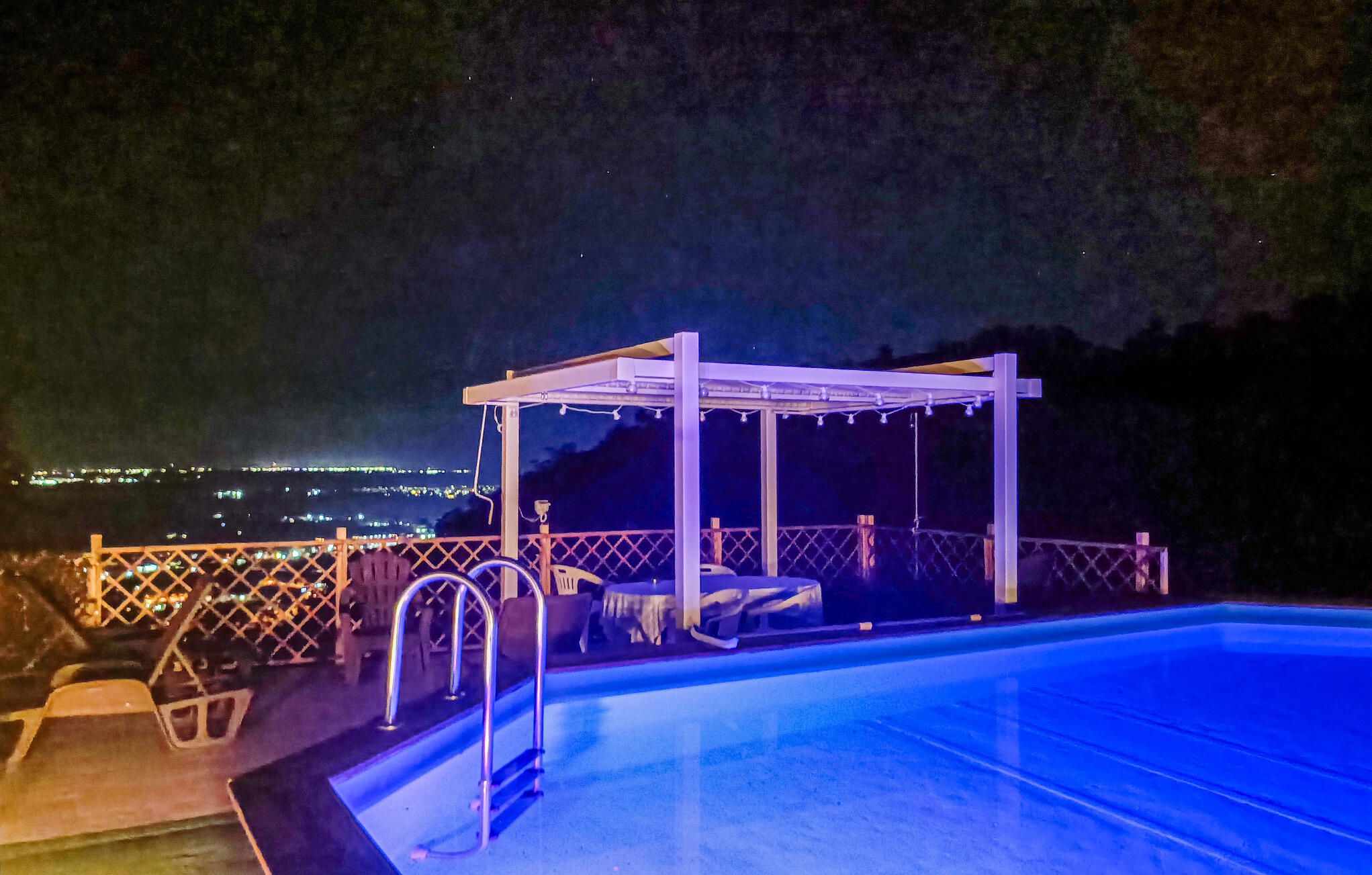 A swimming pool with a pergola structure, a ladder, and a view of a cityscape at night.
