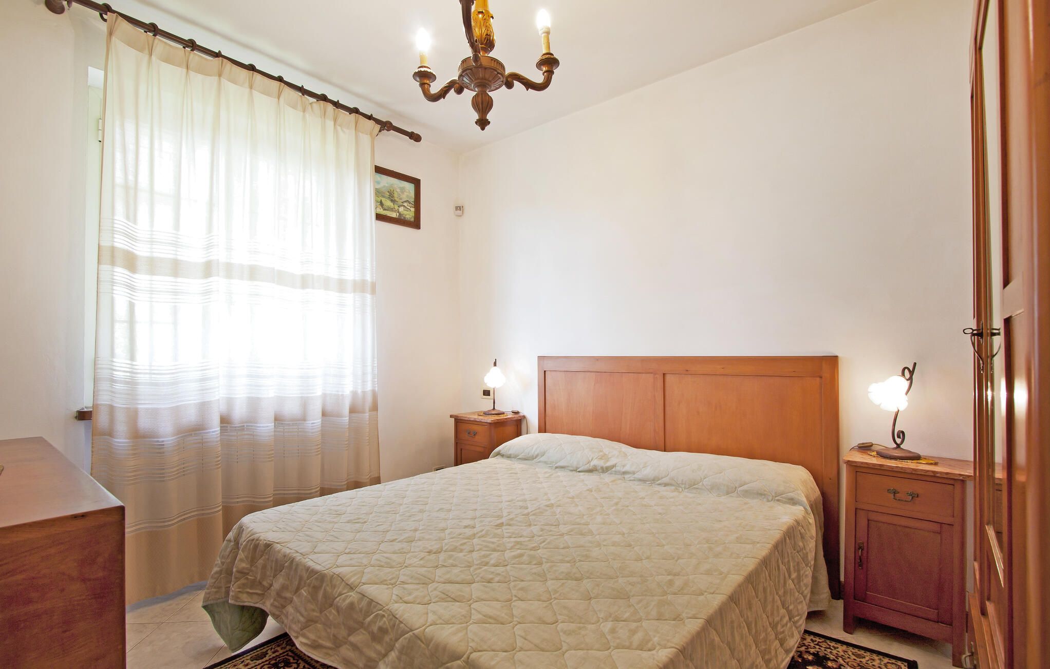 Ein Schlafzimmer mit Bett, Nachttisch, Kommode, einer Kronleuchter und einem Fenster mit Vorhängen.