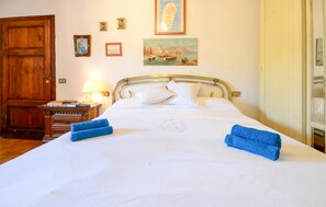 Cama bem arrumada com almofadas e toalhas dobradas, mesa de cabeceira de madeira com abajur, e uma porta à esquerda.