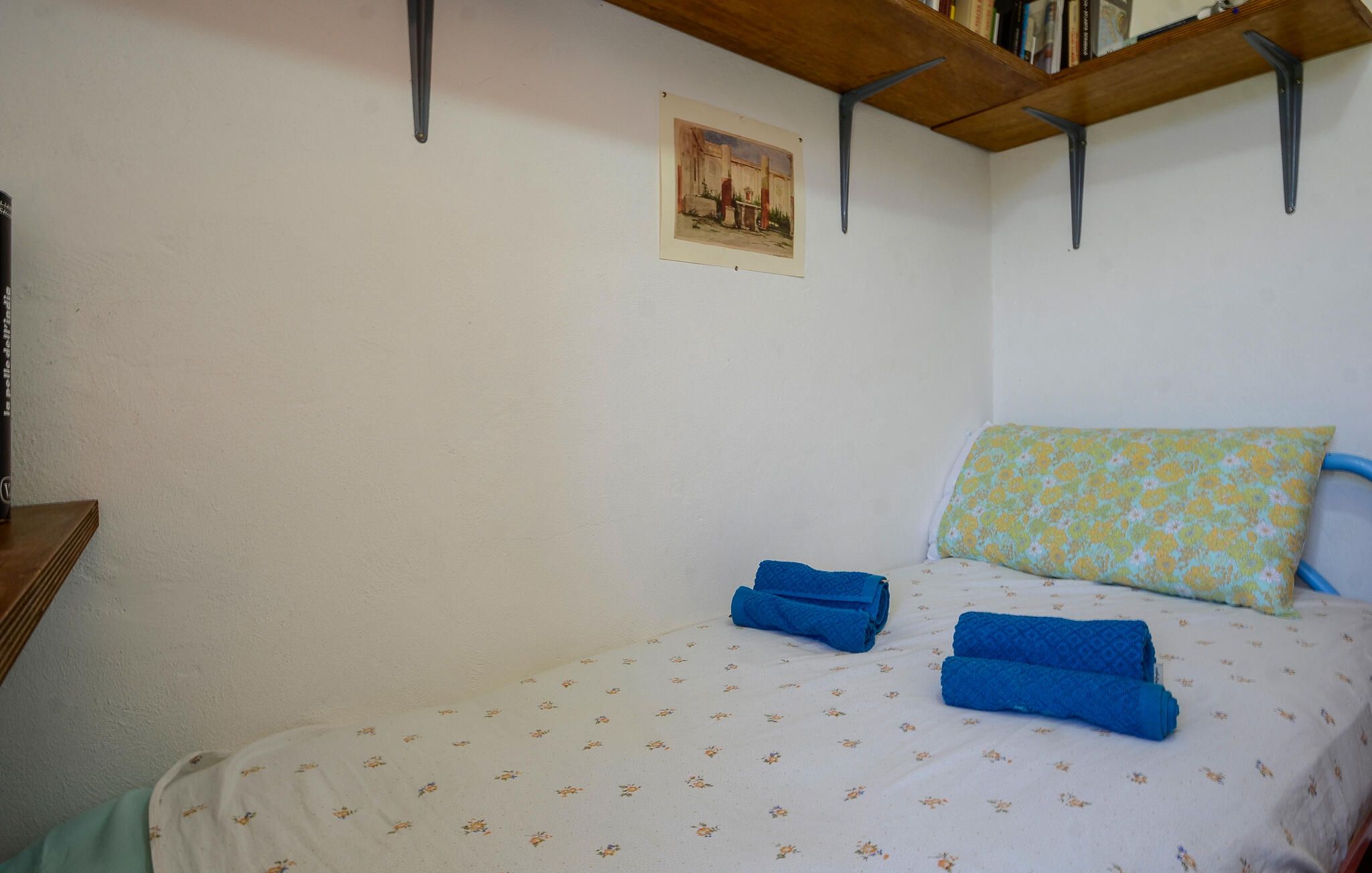A bedroom with a bed, pillows, a painting, and a shelf with books.