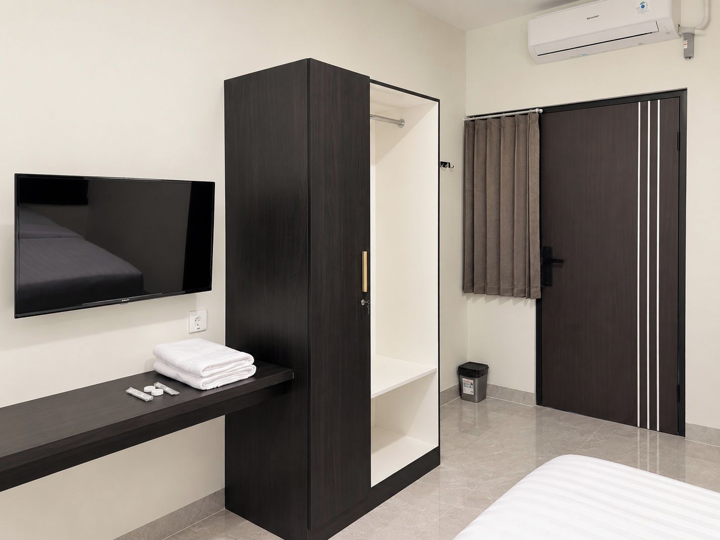 Habitació d'hotel moderna amb un televisió de pantalla plana penjada a la paret, un armari de fusta fosca, una petita taula de nit amb una tovallola i un llit amb matalàs blanc.