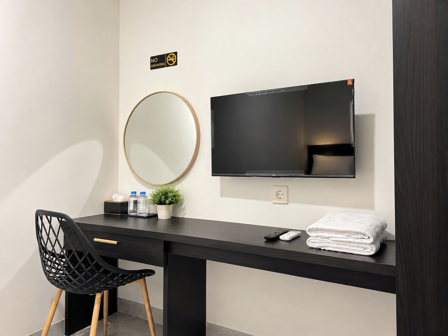 A hotel room with a black desk, a round mirror, a flat-screen TV, a chair, a small plant, and a stack of towels.