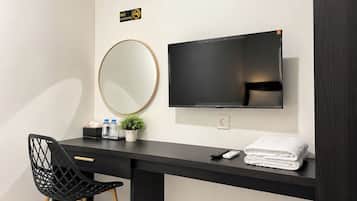 A hotel room with a black desk, a round mirror, a flat-screen TV, a chair, a small plant, and a stack of towels.