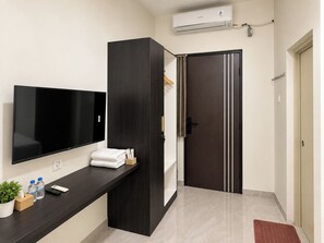 Habitación de hotel con televisor de pantalla plana, armario de madera oscura, puerta con una toalla colgada, una planta pequeña y una alfombra roja.