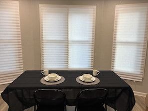 A dining area with a table set for two, two white cups, and two white bowls on placemats, flanked by two windows with blinds. - Pro Cleaned Home for Working Professionals (Kenosha)