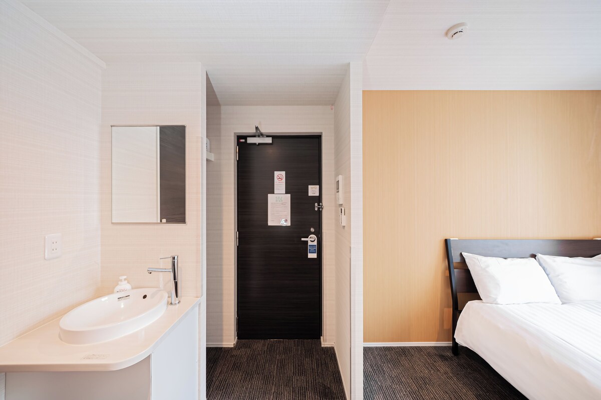 A hotel room with a white sink, a mirror, a dark door, and a bed with white bedding.