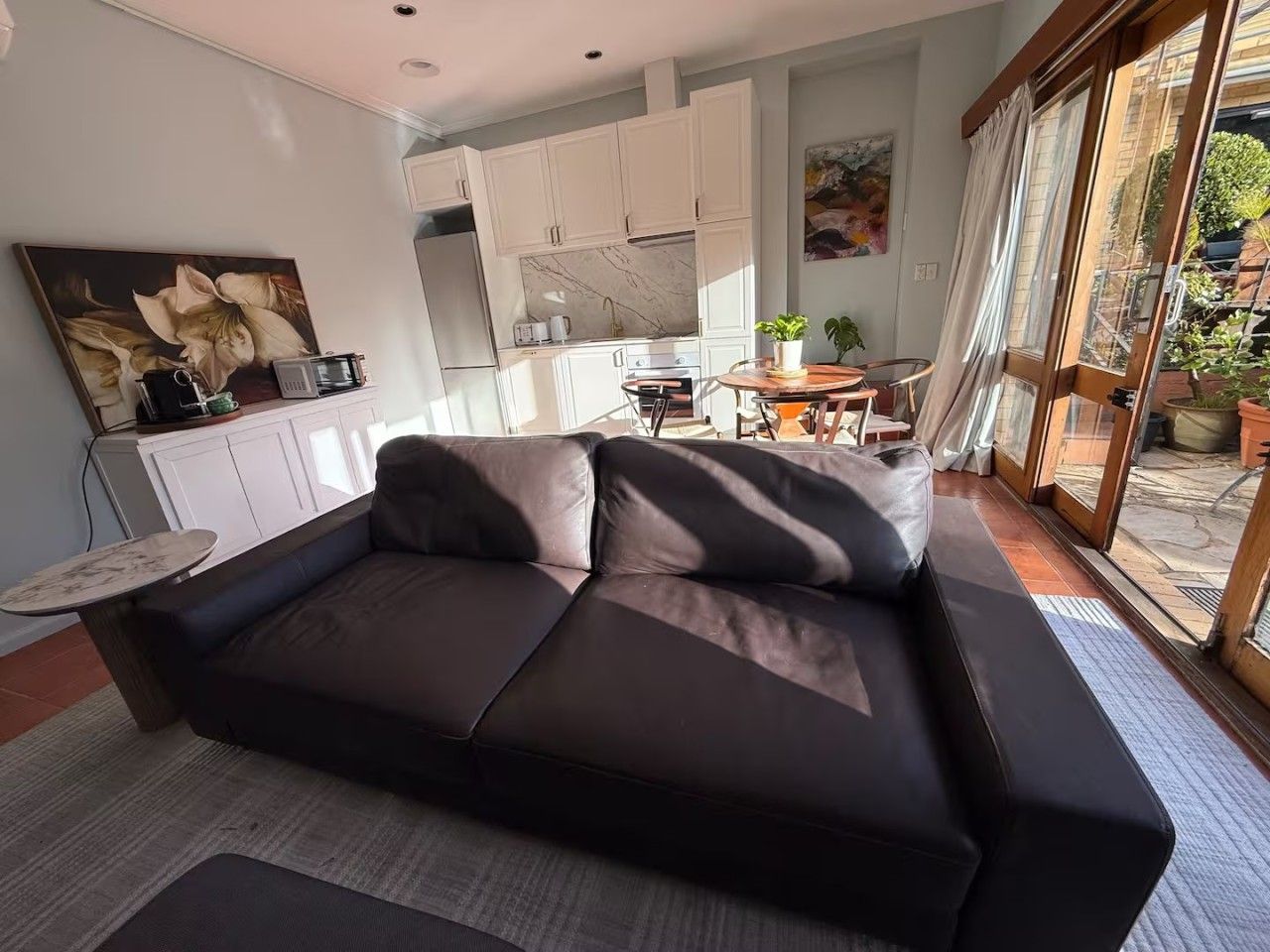 En moderne stue med en mørk lædersofa, et lille rundt bord og et køkken i baggrunden.