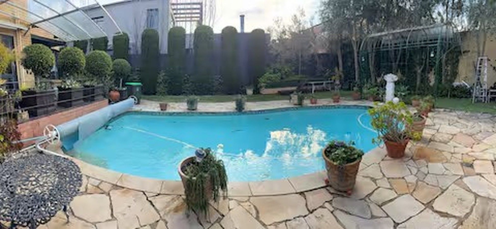 Una piscina con un patio de piedra, rodeada de plantas en macetas y una zona cubierta.