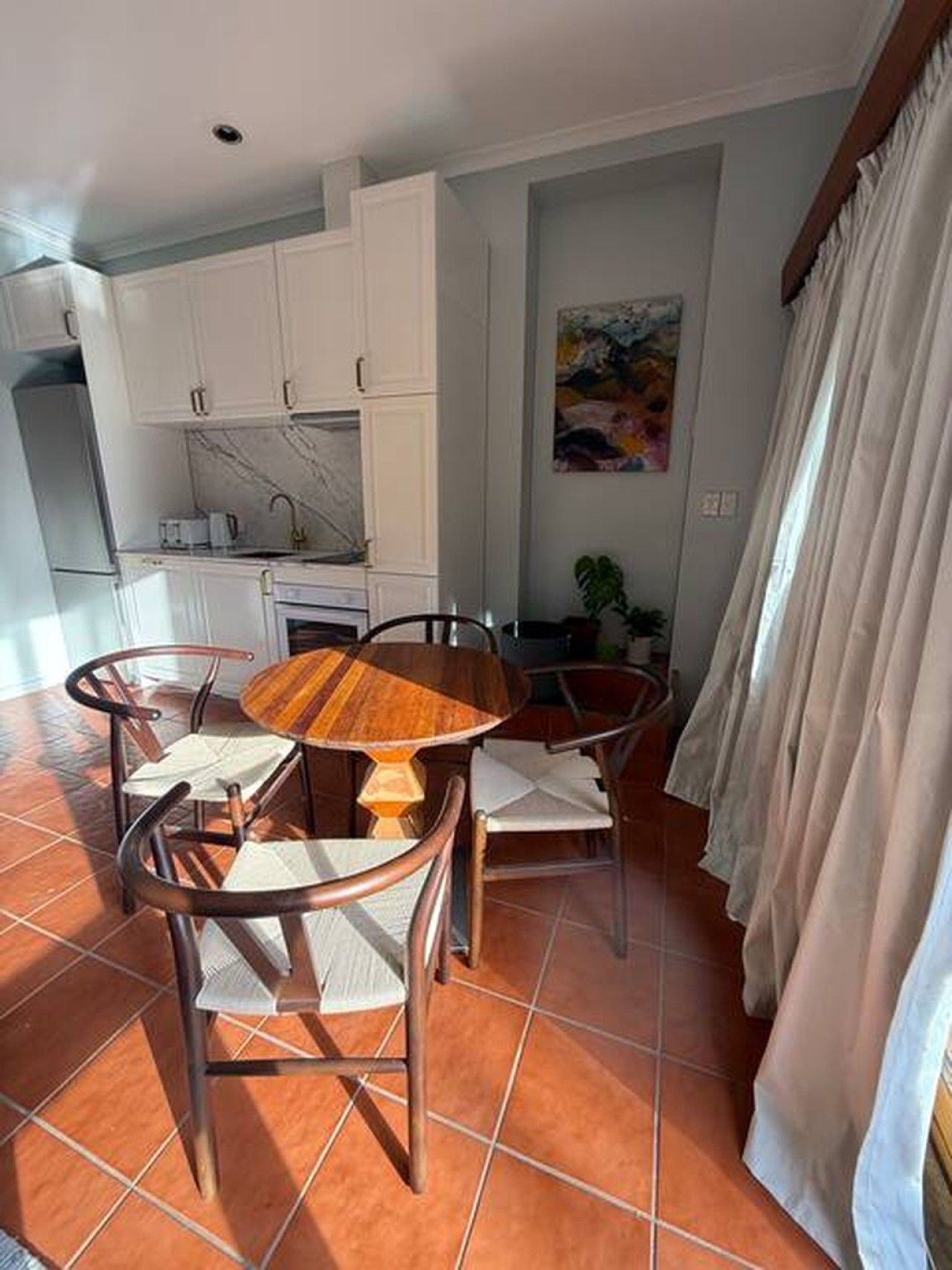 Uma sala de jantar com uma mesa de madeira redonda e quatro cadeiras, uma cozinha com armários brancos e um quadro na parede.