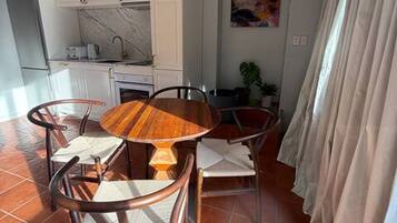 Uma sala de jantar com uma mesa de madeira redonda e quatro cadeiras, uma cozinha com armários brancos e um quadro na parede.