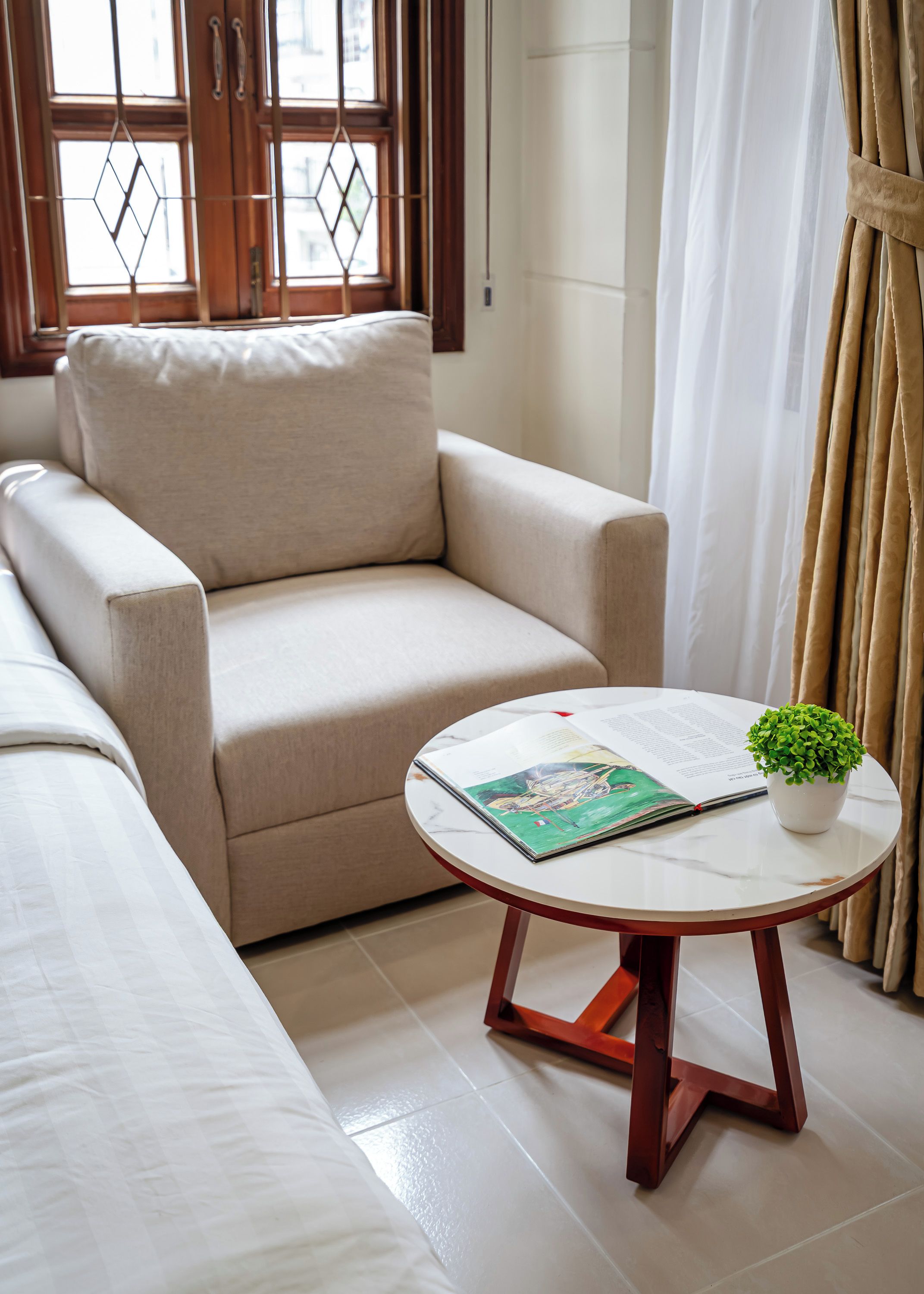 A living room with a beige sofa, a round coffee table with a book and a plant, and a window with curtains.