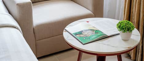 Uma sala de estar com um sofá bege, uma mesa de centro redonda com um livro e uma planta, e uma janela com cortinas.