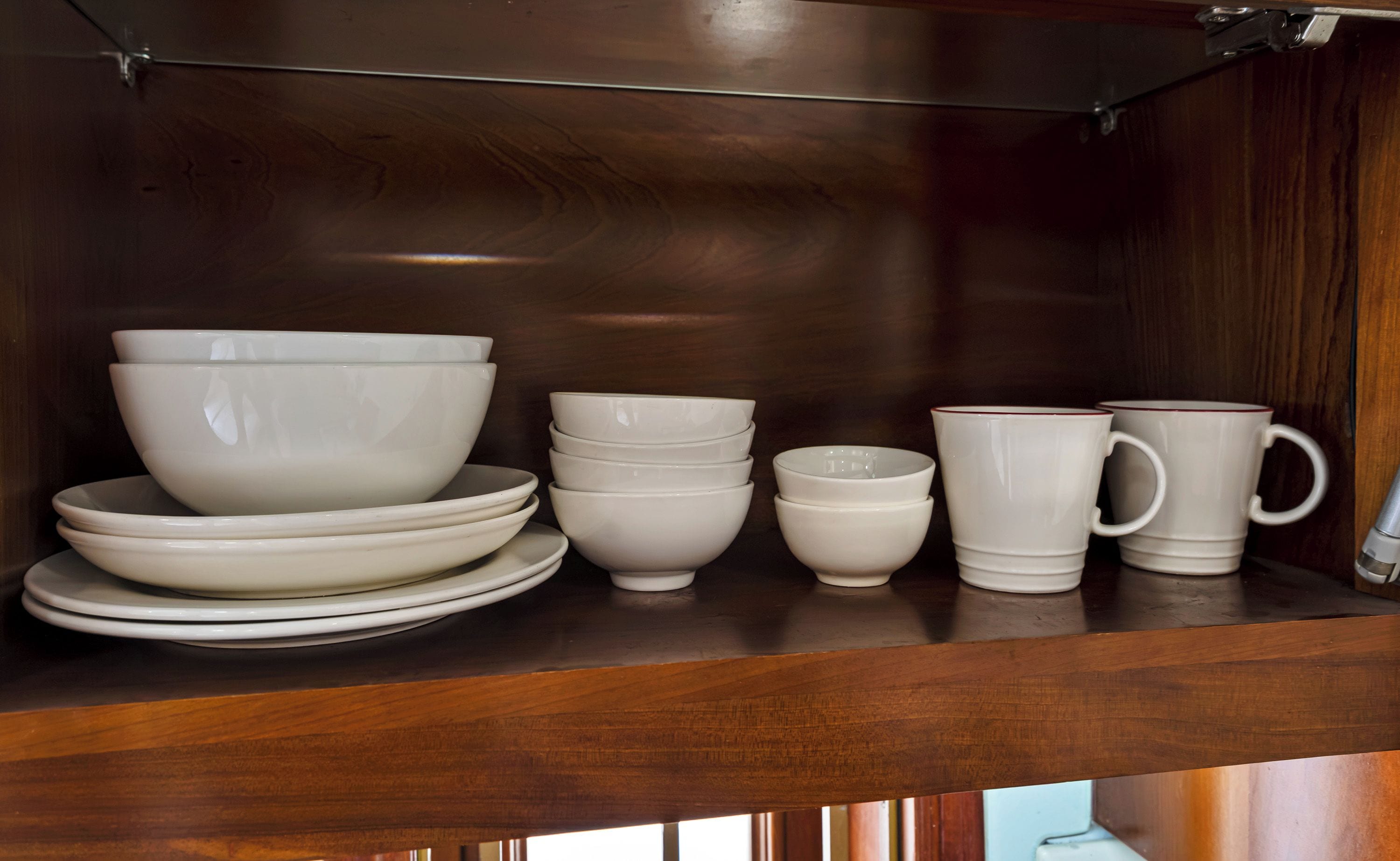 A wooden cabinet with a glass shelf holding white ceramic dishes and cups.