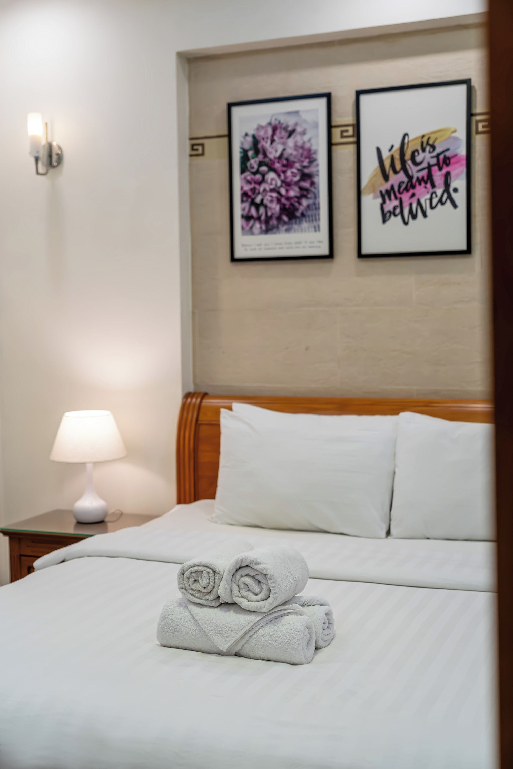 A neatly made bed with rolled towels, a bedside lamp, and two framed pictures on the wall.