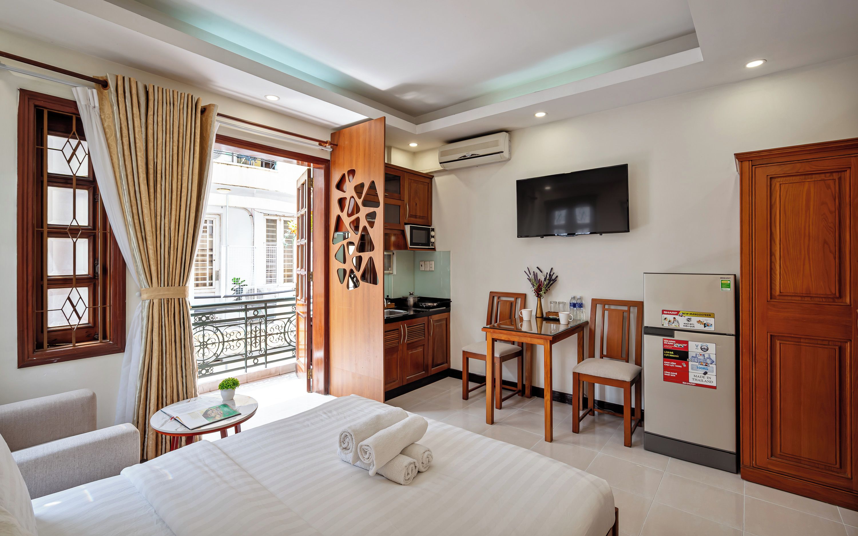 A hotel room with a bed, a dining table with chairs, a kitchen area, and a TV mounted on the wall.