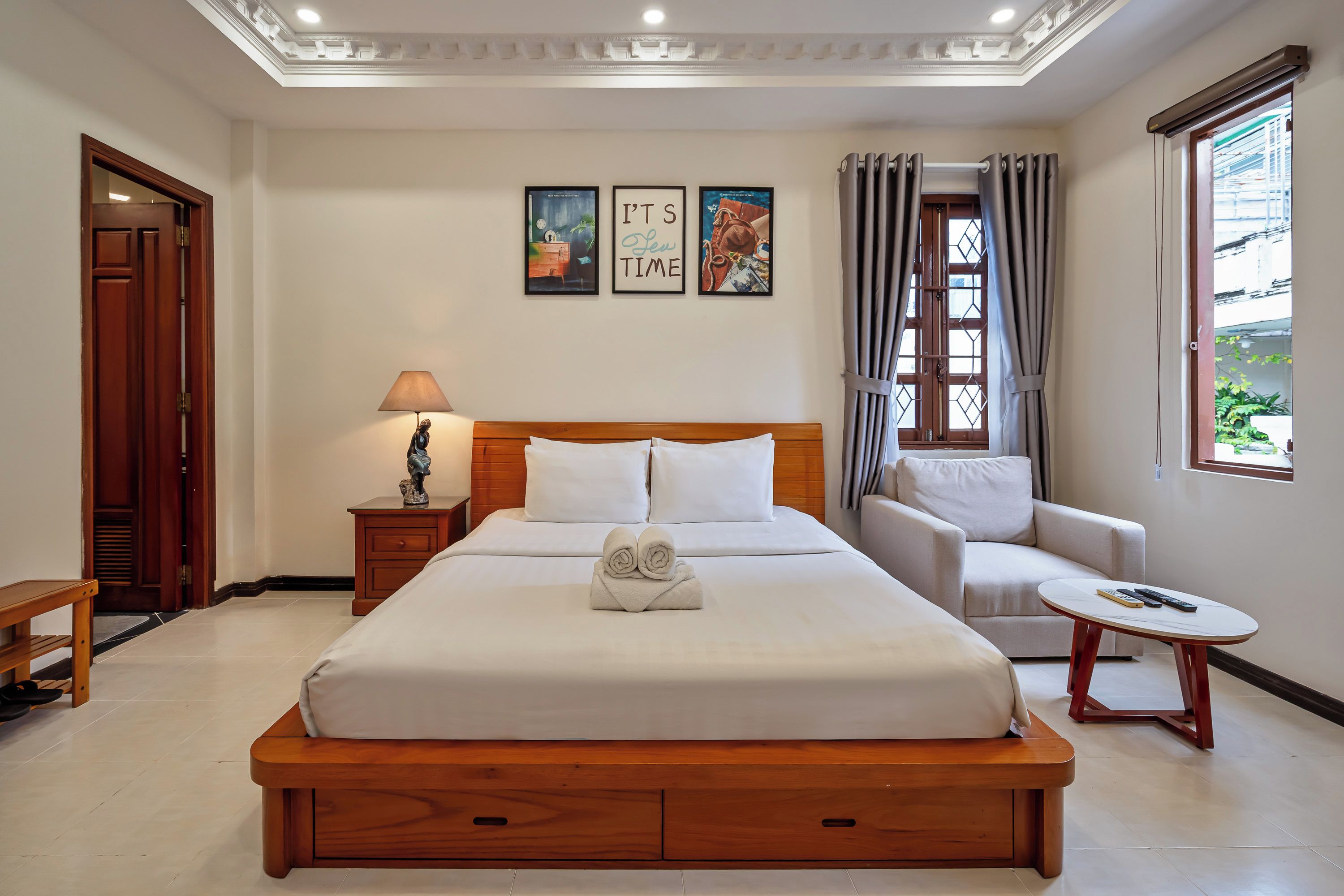 A hotel room with a large bed, a bedside table with a lamp, a chair, a small table, and a window with curtains.