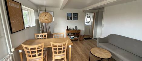 A dining area with a wooden table and chairs, a hanging light fixture, and a grey sofa.
