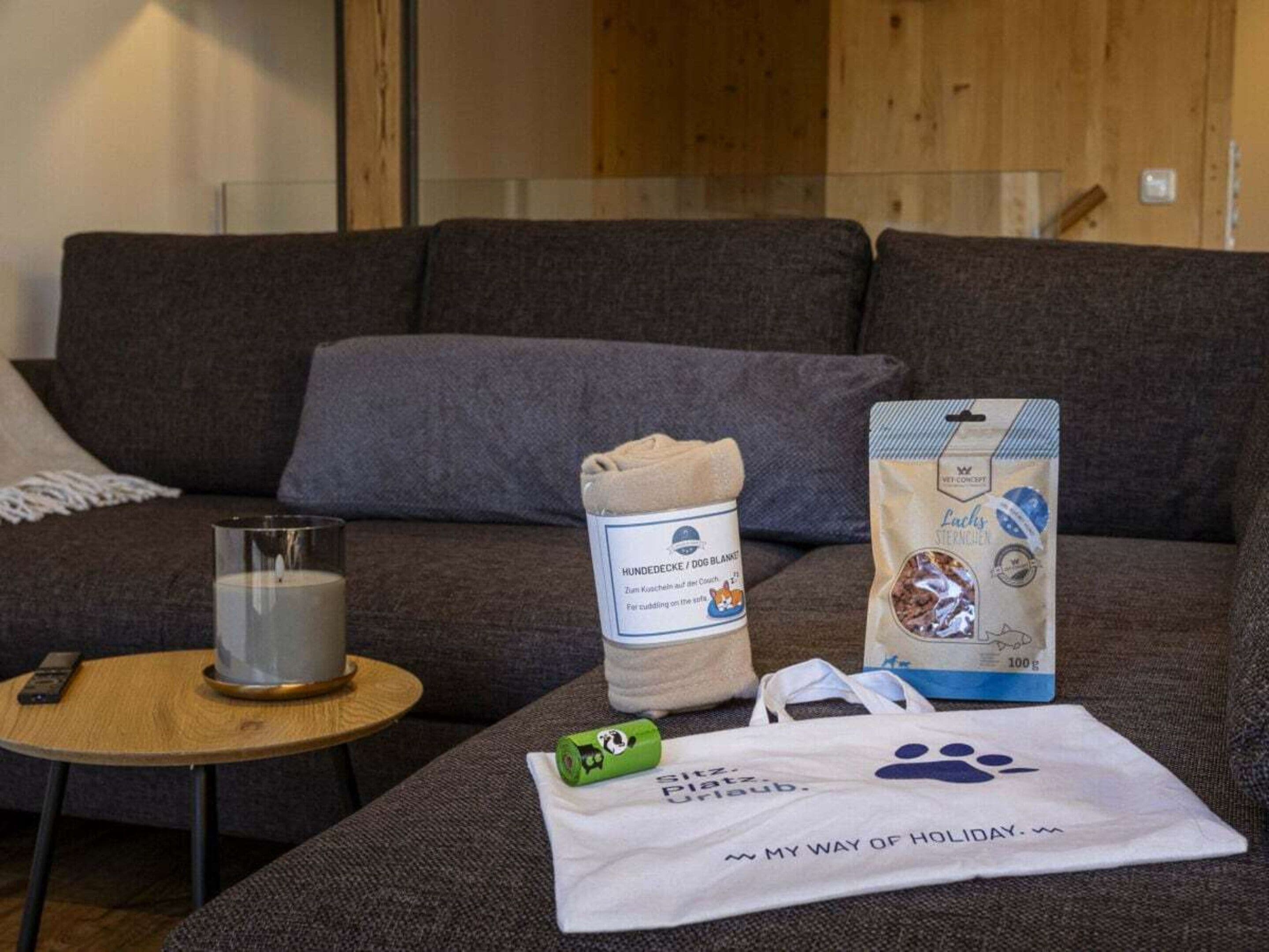 A living room with a grey sofa, a glass candle holder, a rolled-up blanket, a pet food bag, and a tote bag with a paw print.
