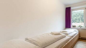 Una cama individual con ropa de cama blanca en una habitación con paredes blancas y una ventana con cortinas moradas.