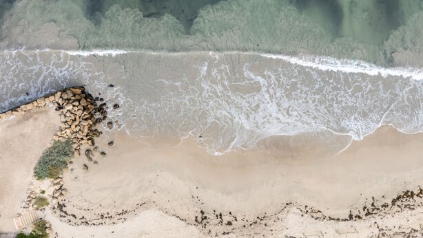 Vista aérea de uma praia com costa rochosa, ondas e uma área arborizada ao fundo.