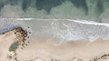 An aerial view of a beach with a rocky shoreline, waves, and a forested area in the background.