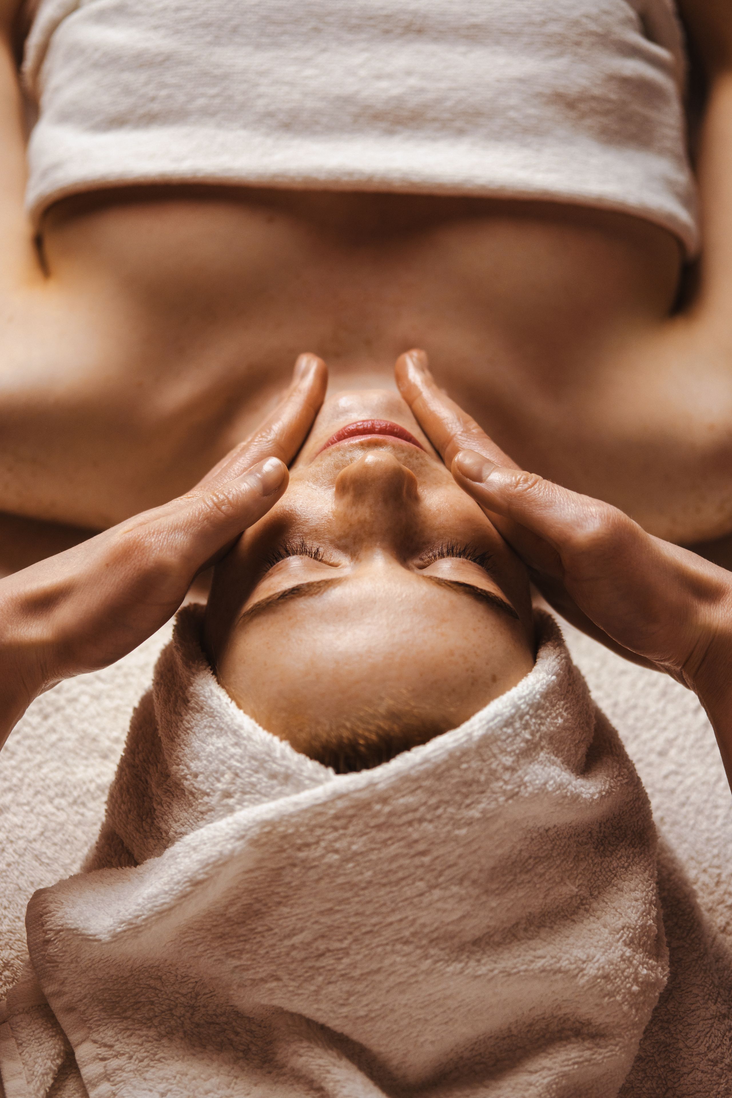 A person receiving a facial treatment with hands on their face, wrapped in a towel.