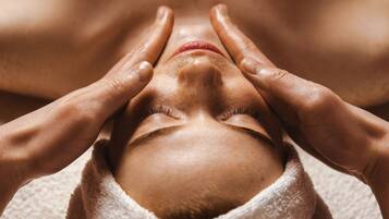 A person receiving a facial treatment with hands on their face, wrapped in a towel.