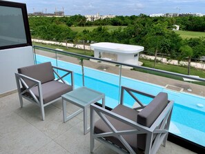 Un balcon avec deux chaises et une petite table, d’où l’on peut admirer une piscine et un paysage verdoyant, avec des bâtiments en arrière-plan.