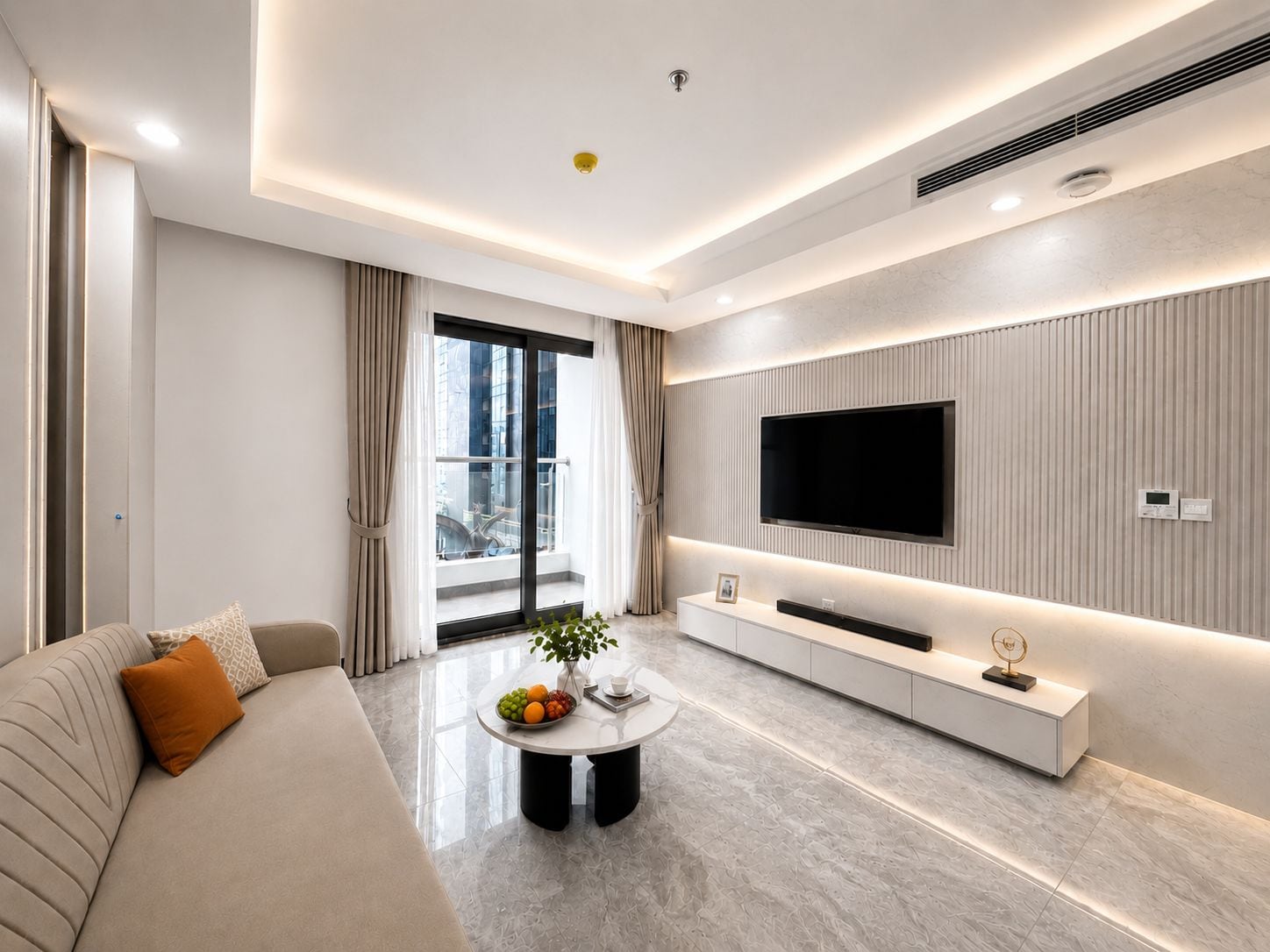A modern living room with a sofa, a coffee table with fruit, a TV mounted on the wall, and a balcony with curtains.