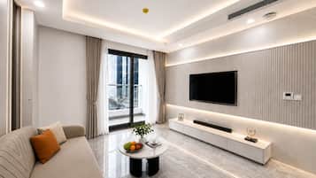 A modern living room with a sofa, a coffee table with fruit, a TV mounted on the wall, and a balcony with curtains.