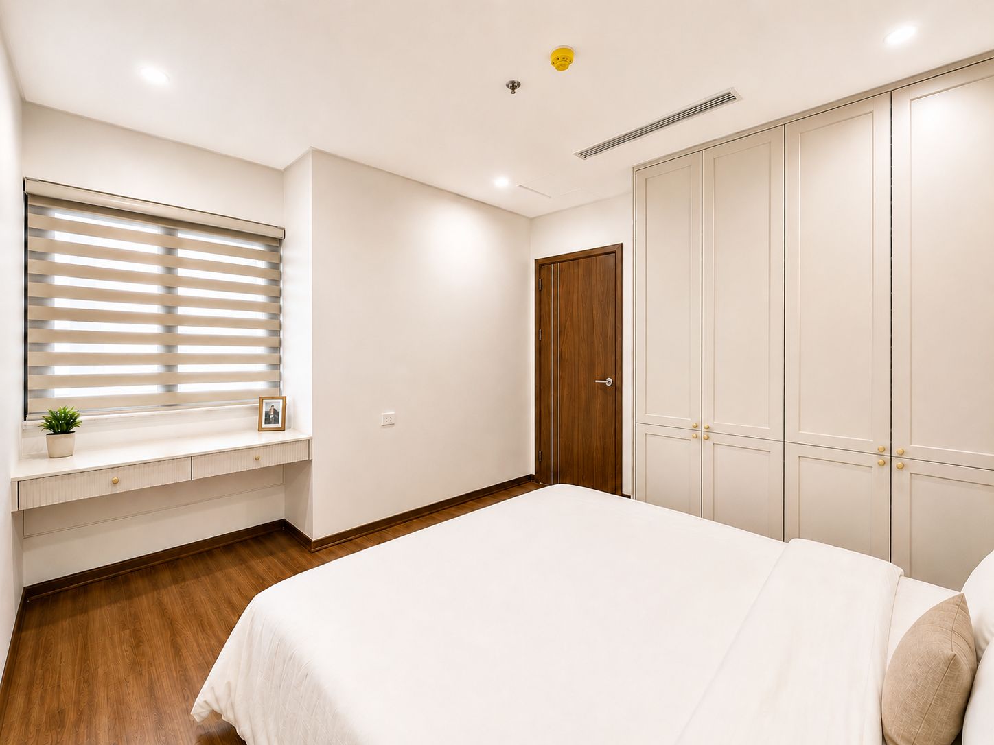 A bedroom with a bed, a window with blinds, a bedside table with a plant, and a wardrobe.