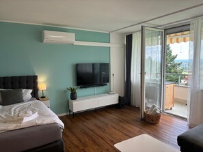 A modern bedroom with a bed, a TV, a white console, a plant, and a balcony with a view. - Modern vacation apartment with panoramic views and a cozy, comfortable atmosphere (Oberteuringen)