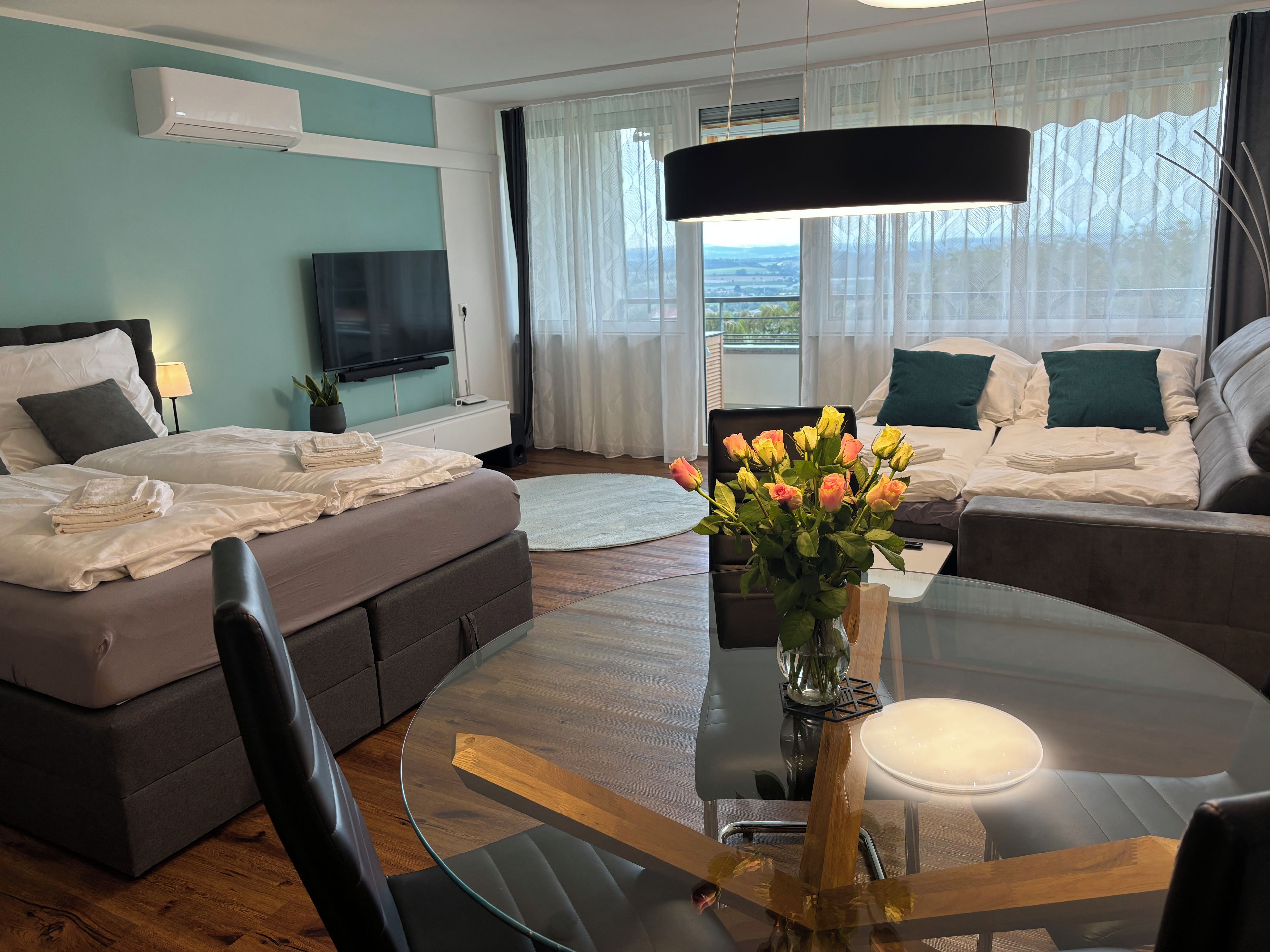 Ein modernes Hotelzimmer mit Schlafsofa, Flachbildfernseher, Esstisch mit Stühlen und Blick ins Freie durch große Fenster.